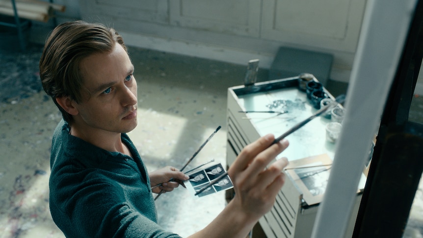 Colour still of Tom Schilling painting with brush in right hand and holding photo in left hand in 2018 film Never Look Away.