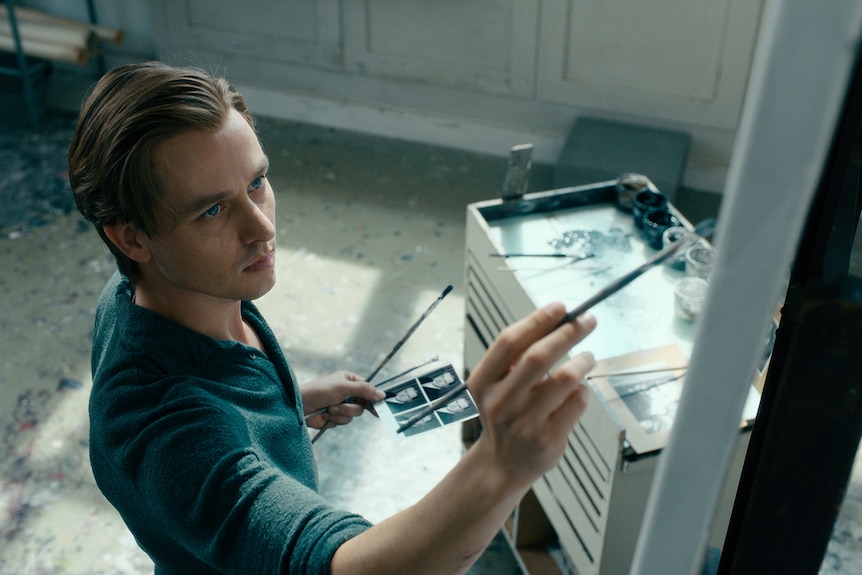 Colour still of Tom Schilling painting with brush in right hand and holding photo in left hand in 2018 film Never Look Away.