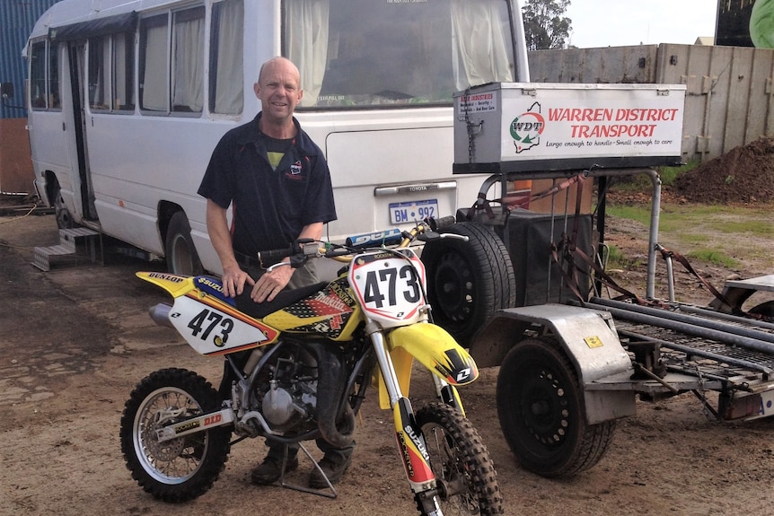 Bald man standing next to a yellow dirt bike with number 473, in front of white bus.