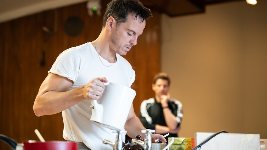 Andrew Scott at a sink with a kettle in one hand and a cigarette in the other. Sam Yates, out of focus, looks on behind him.