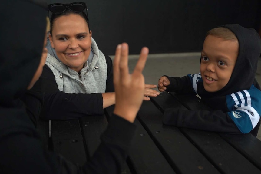 A woman smiles as she sits next to her son who has dwarfism. Person wearing a black hoodie holds up three fingers in foreground