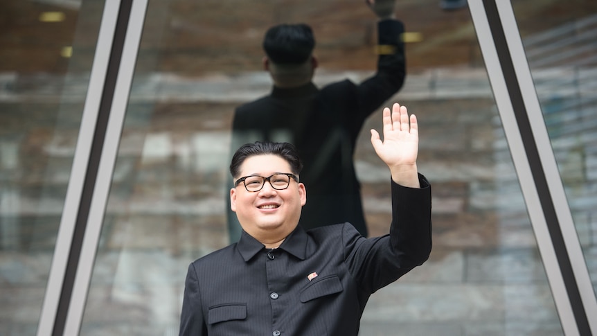 Howard X, dressed as Kim Jong Un in black suit, hair with shaved sides and black-rimmed glasses, smiles and waves.