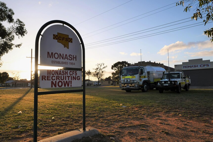 A sign reads Monash established 1954, recruiting now. Below is lush lawn with the sun beaming. Two fire vehicles are behind.