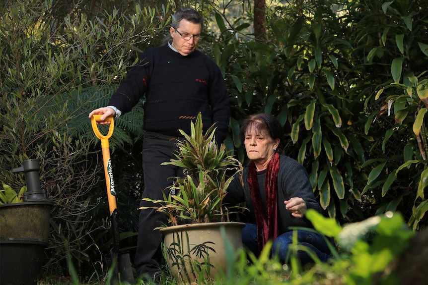 A man stands over a plant with his shovel, while a woman crouches on the ground in front of him.