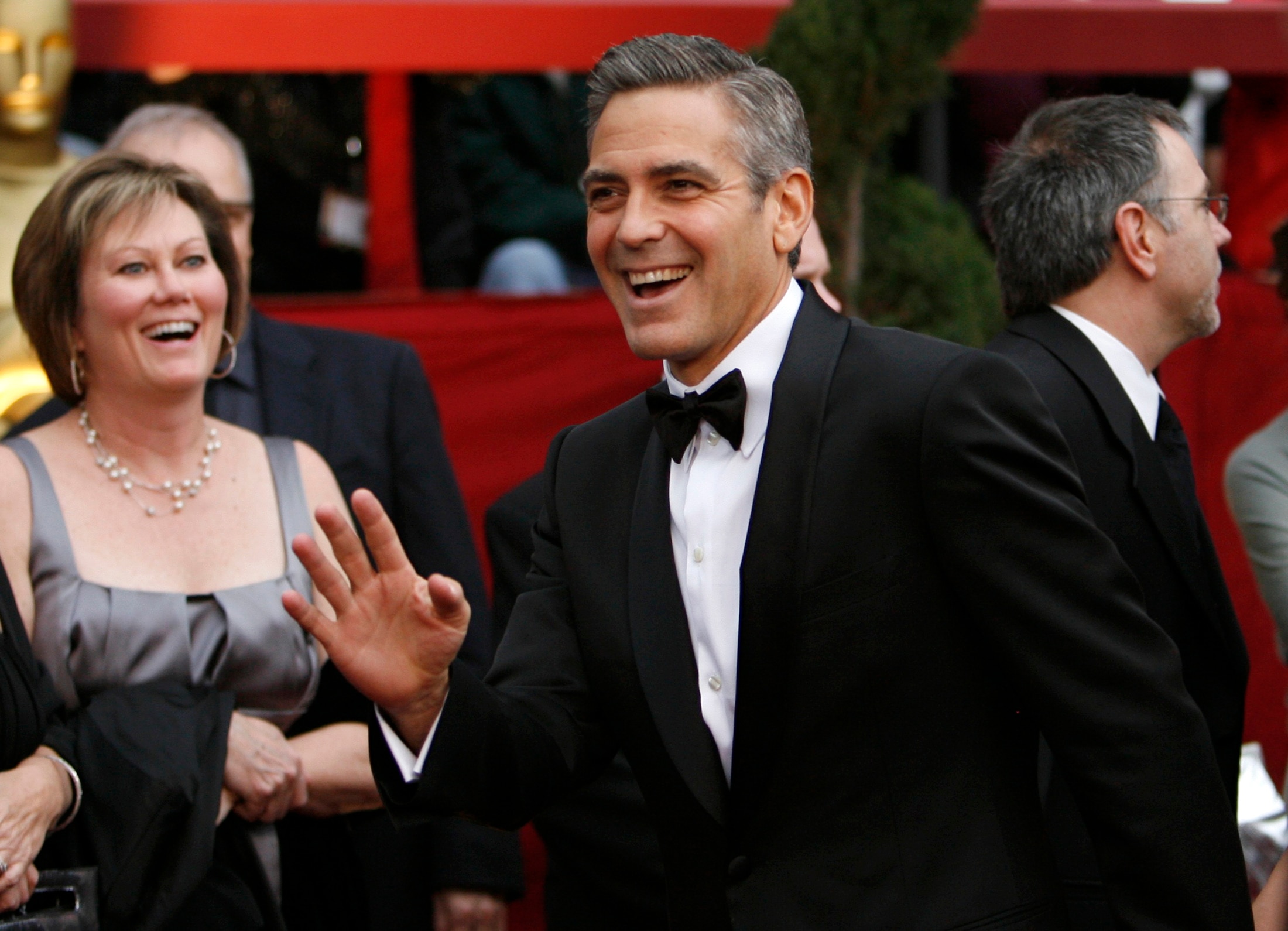 George Clooney in a black suit and bow tie on a red carpet smiling and waving. 