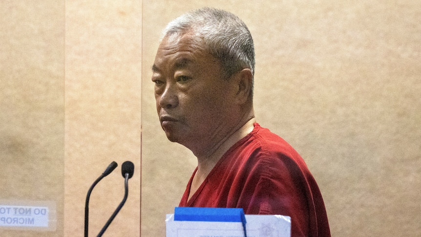 A man in a red jumpsuit appears behind a glass screen. 