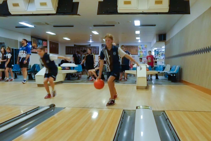 Two students are mid motion bowling, wearing school uniforms. Other students in background at bowling alley