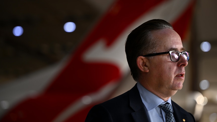 A middle-aged white man in a suit with glasses and slicked-back hair stands in front of the red tail of a Qantas plane.