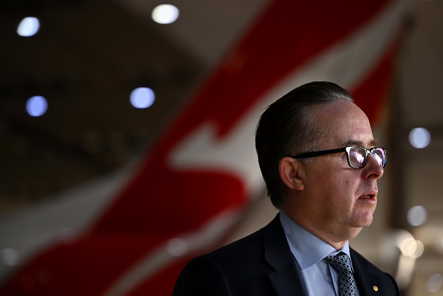 A middle-aged white man in a suit with glasses and slicked-back hair stands in front of the red tail of a Qantas plane.