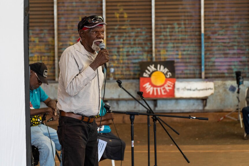 An elderly Indigenous man addressing a crowd with a microphone, with a Blak Lives Matter sign behind him
