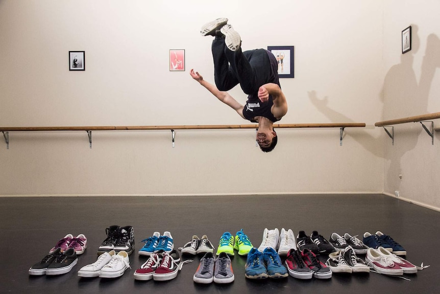 A young man upside down part way through a breakdancing flip in a dance studio with shoes in front