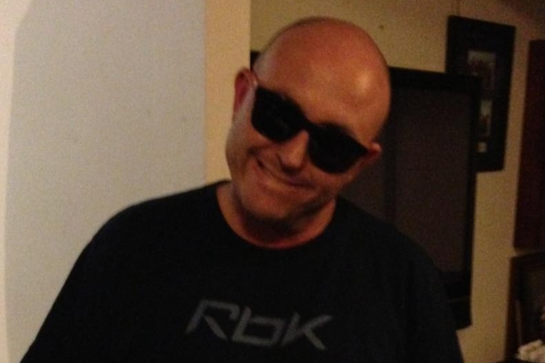 A bald man wearing dark sunglasses smiles and inclines his head.