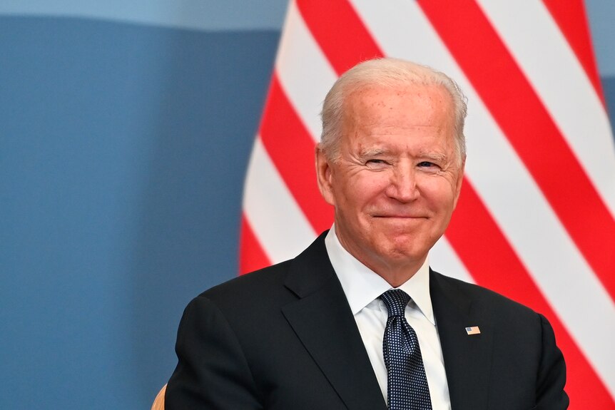 Joe Biden dressed in a dark suit smiles in front of the US flag.
