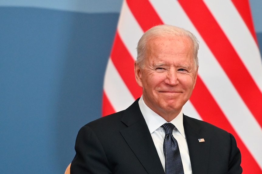 Joe Biden dressed in a dark suit smiles in front of the US flag.