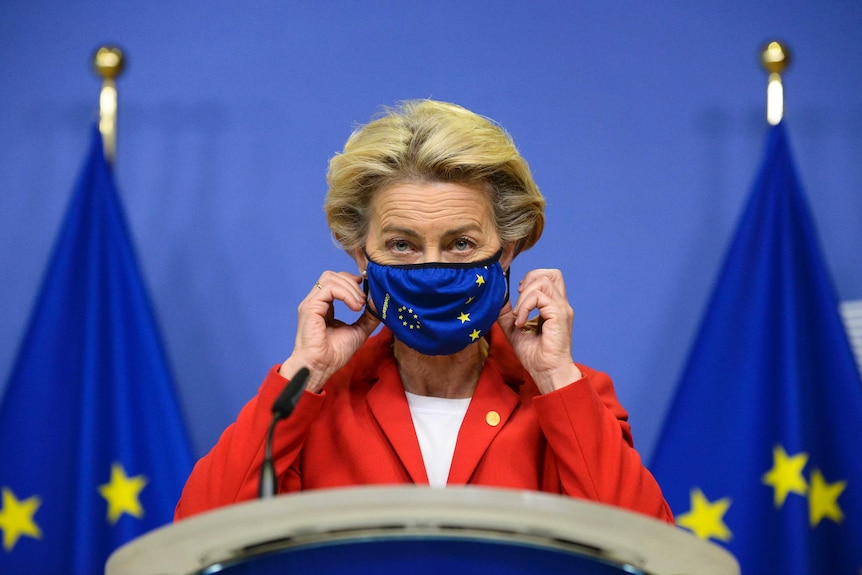 A blonde woman adjust the straps of her EU flag face mask while standing behind a podium.