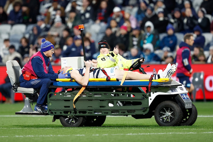 A Geelong AFL player puts his thumbs up as he lies on a mobile stretcher with a neckbrace and a trainer supporting his head.