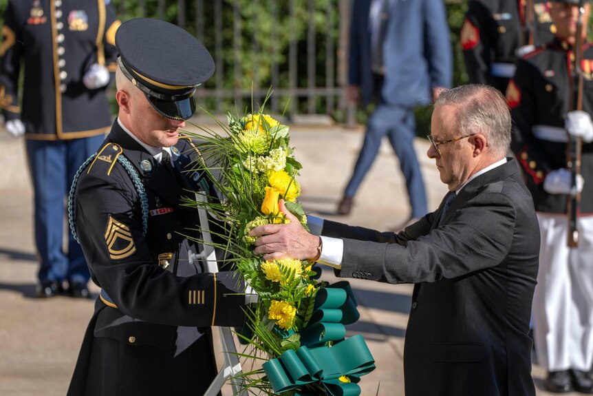 Anthony Albanese hands a uniformed officer a large wreath with yellow Australian flowers on it.