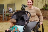 Molly is smiling and has both hands on her wheelchair's wheels. Her son is in a pram attached to the front of her chair.