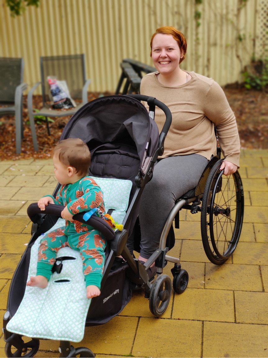 Molly is smiling and has both hands on her wheelchair's wheels. Her son is in a pram attached to the front of her chair.