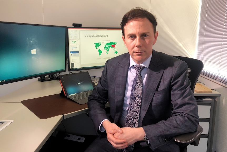 A man in a suit sits in front of a computer in an office and looks at the camera.