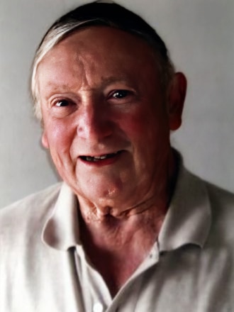 An elderly man with white hair and dark eyes in a white polo t-shirt smiles. 