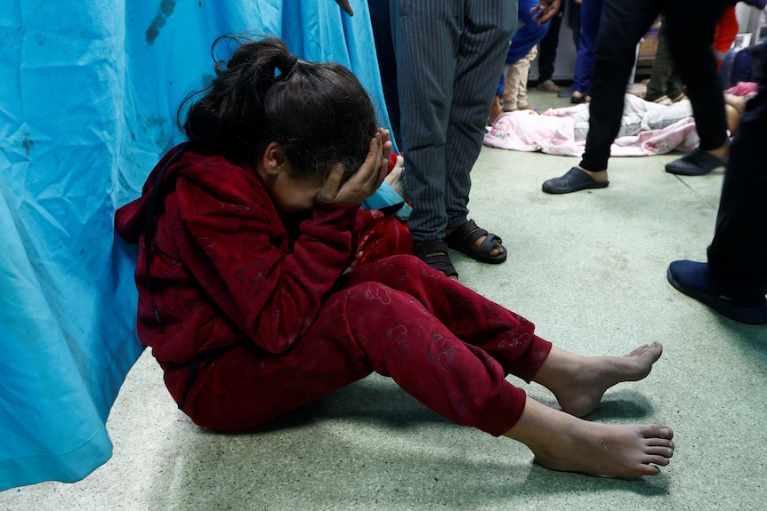 A girl, around 6 yrs old, barefoot, cries with her head in her hands on a hospital floor.