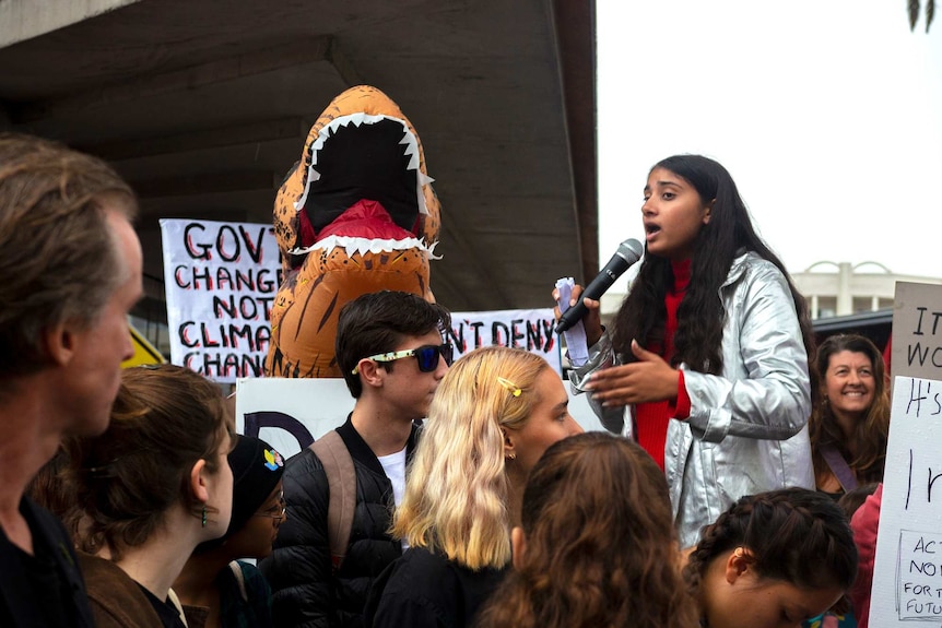 A woman of South Asian descent is standing above a crowd and speaking into a mic while wearing a bright silver jacket.