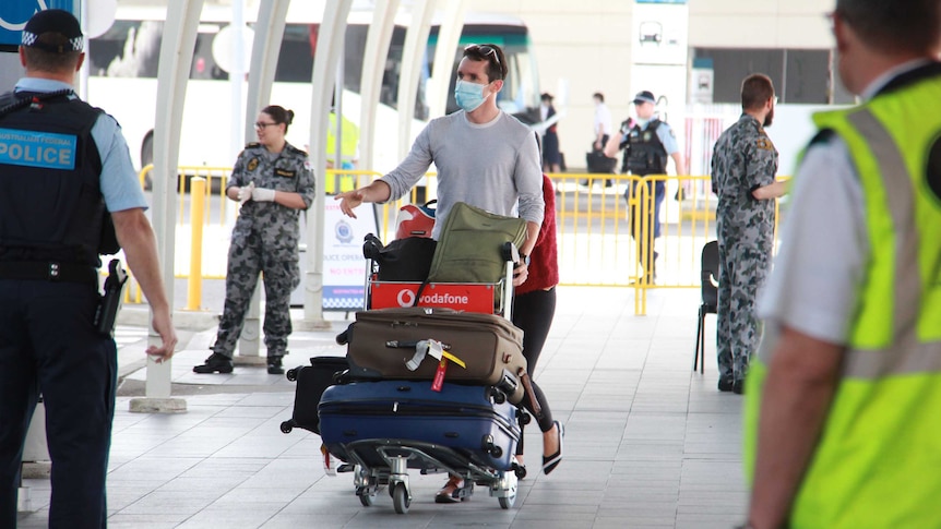 A man wearing a blue surgical mask with a trolley of bags at an arrival hall of an airport.