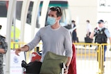 A man wearing a blue surgical mask with a trolley of bags at an arrival hall of an airport.