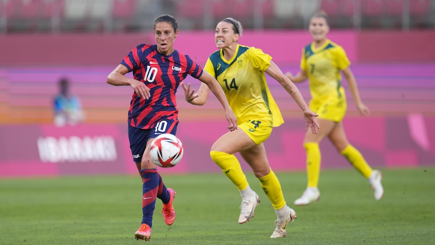 USWNT player Carli Lloyd runs away from Australian Matildas defender Alanna Kennedy, who has a strained look on her face.