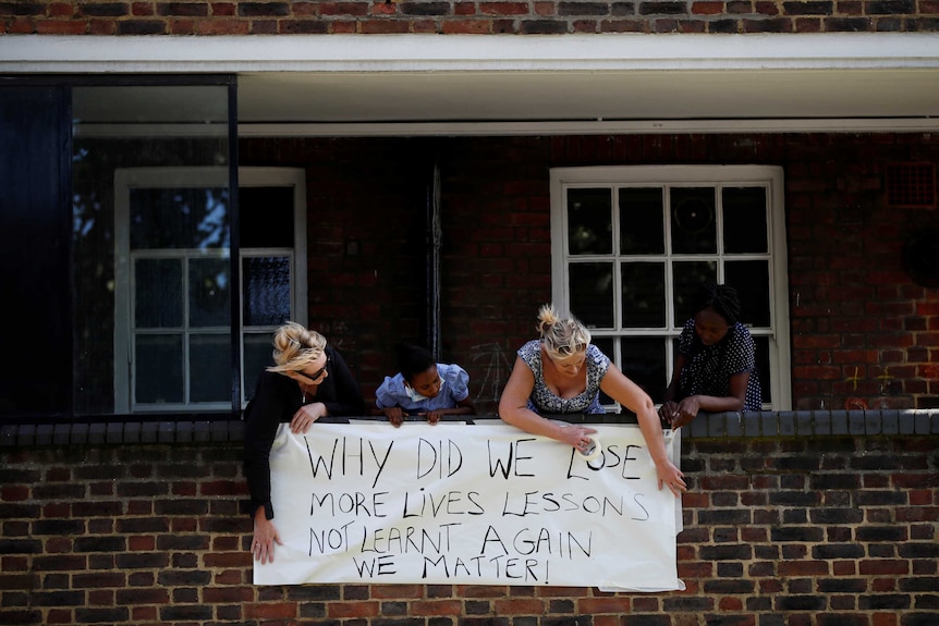 Three residents hang a banner over a brick balcony reading "Why did we lose more lives lessons not learnt again we matter"