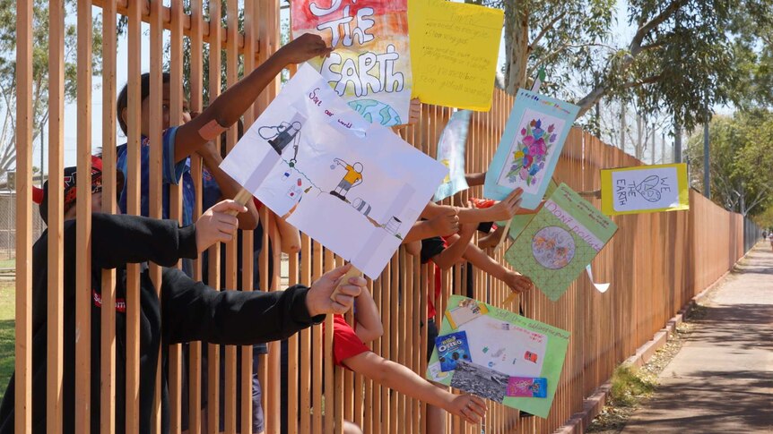Students in Tennant Creek wave signs at passing cars through the schoolyard fence.