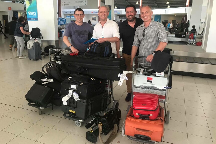 Four men standing at airport terminal with trolley packed with equipment