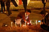 Man lights candle on ground while another man films it on his phone, while flag flying in background.