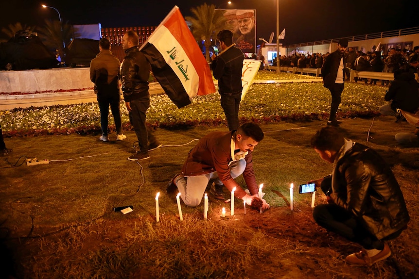 Man lights candle on ground while another man films it on his phone, while flag flying in background.