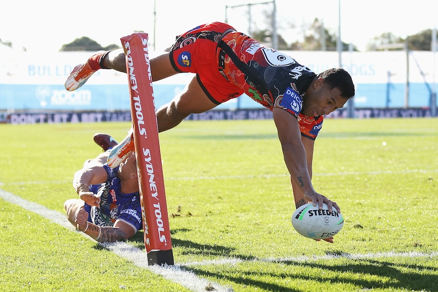 Tautau Moga dives in for a try with the ball in one hand for the Dragons. A Bulldogs defender is on the ground behind him.
