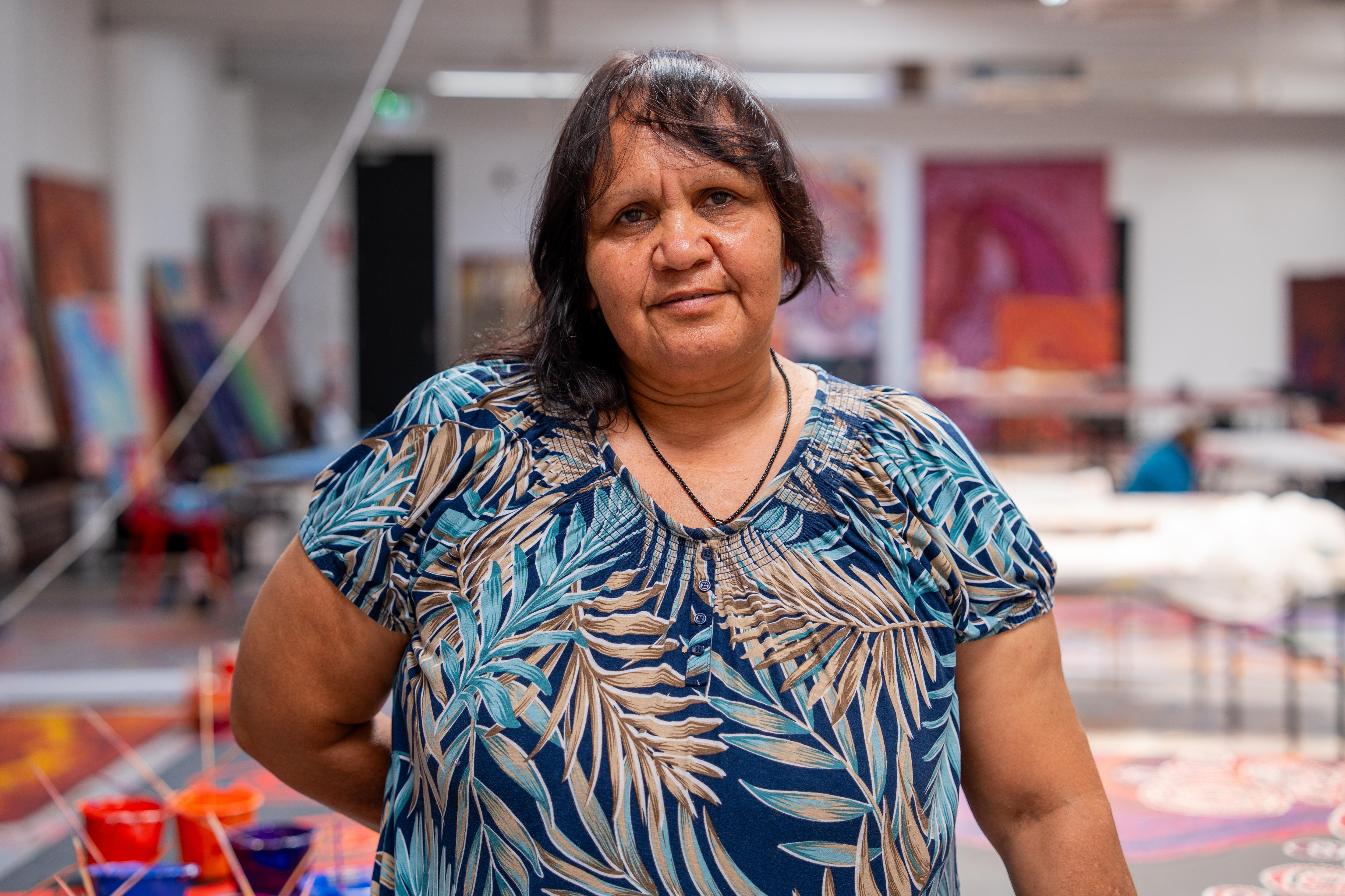 Uma mulher de cabelos escuros e uma blusa colorida está em um estúdio de arte.