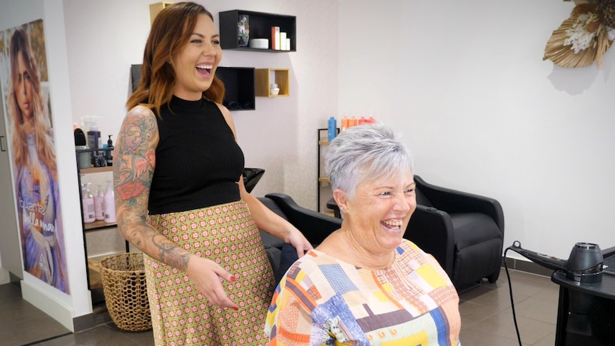 A woman with short hair sits down at a salon chair, a woman stands behind her with her hand slightly stretched out laughing