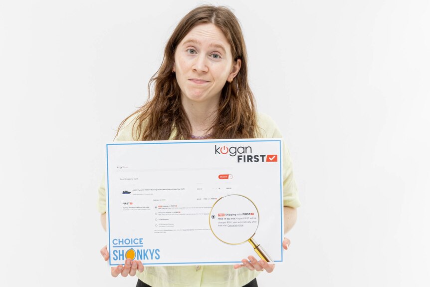 A woman standing in a white room holding a Shonky award with the Kogan logo, and shrugging