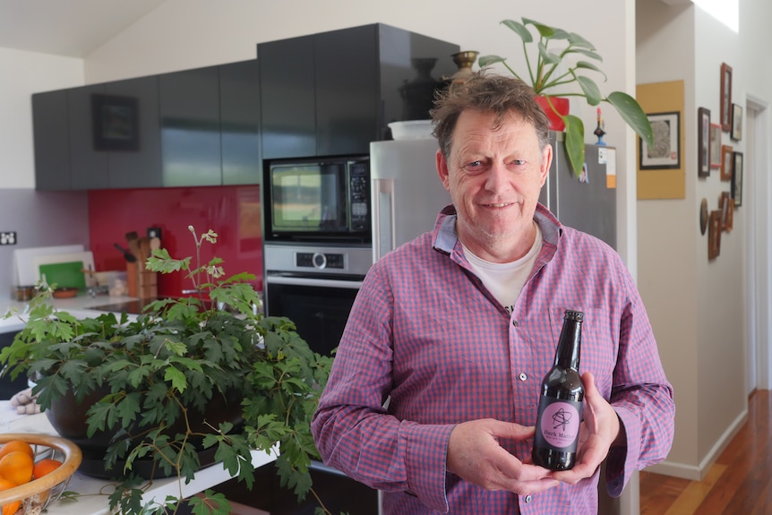 A smiling man in a light purple collared shirt holding an unopened bottle of beer.