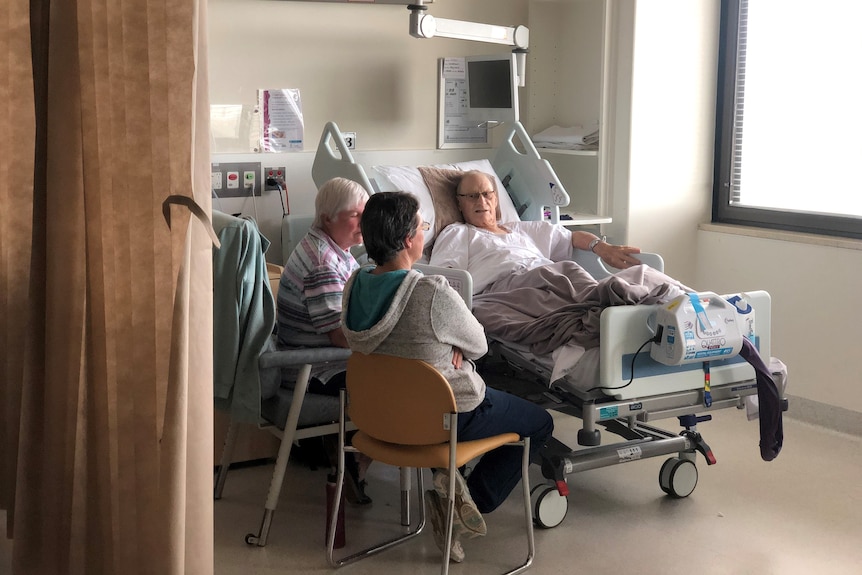 A man in a hospital bed speaks to two women sitting beside him