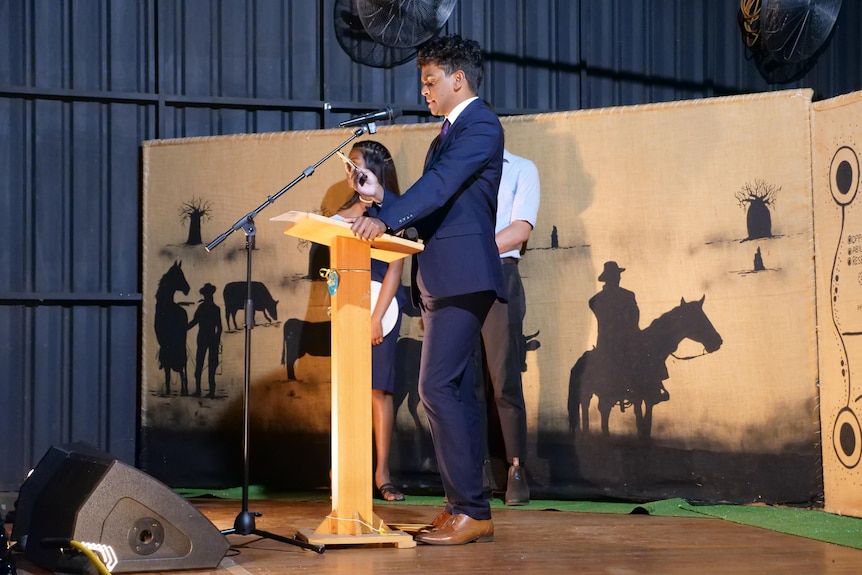 A young Indigenous man wears a suit as he delivers a speech at a lecturn