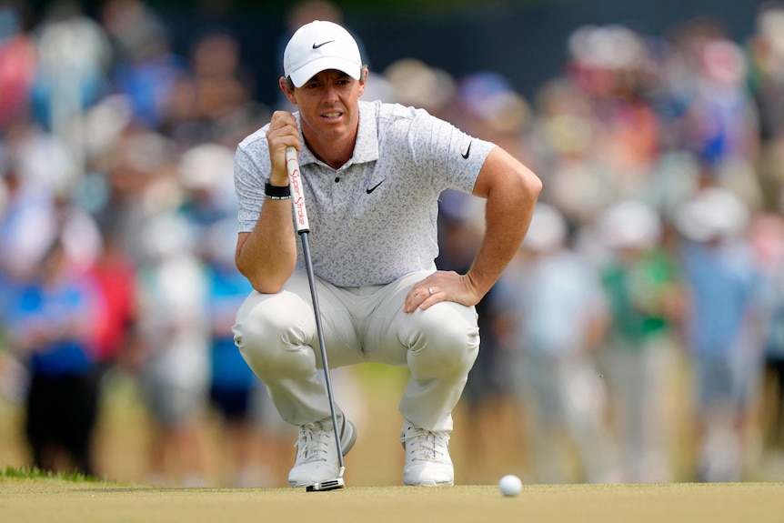A young white man in grey golf clothing man crouches down as he lines up a putt in front of a crowd.