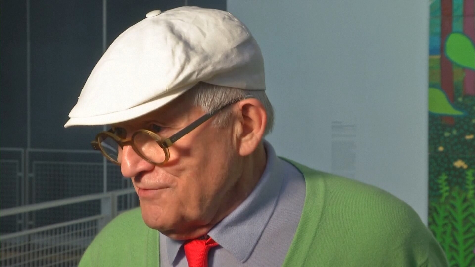 David Hockney in 2017, wearing a hat and signature thick rimmed spectacles.