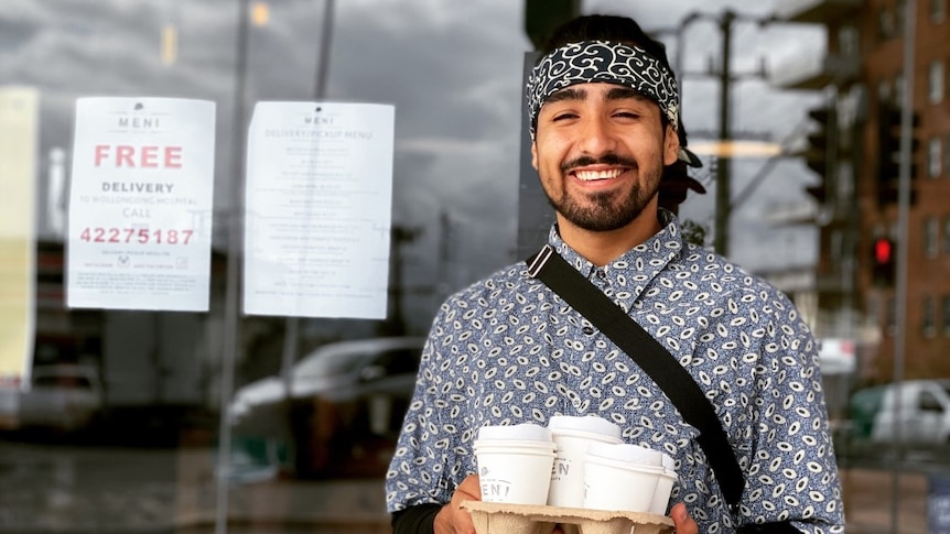 Brandon Jaque holds four coffees while smiling out the front of Meni cafe in Wollongong.