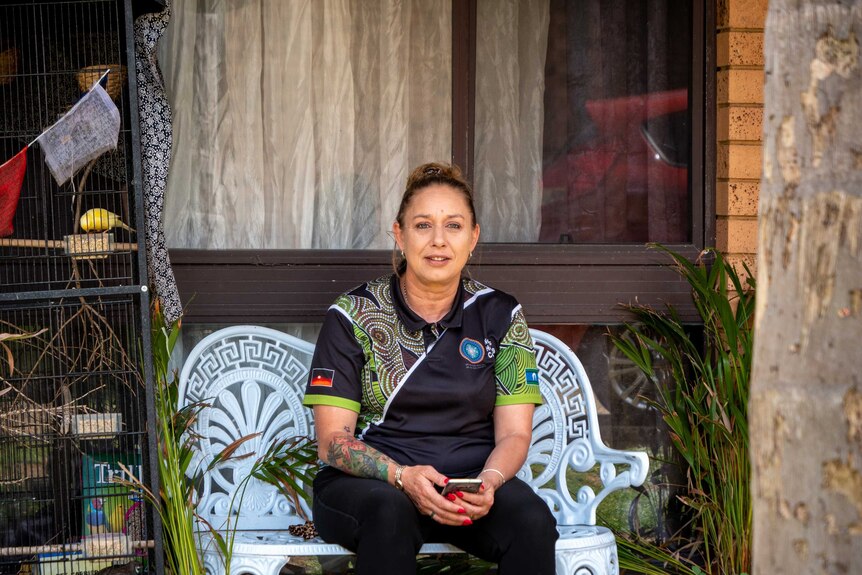 Helen Eason sits on a bench outside a house, she is holding a phone and wearing a polo shirt with Indigenous designs on it.