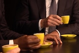 Two men close-up holding small yellow coffee cups with saucers on a wooden table 
