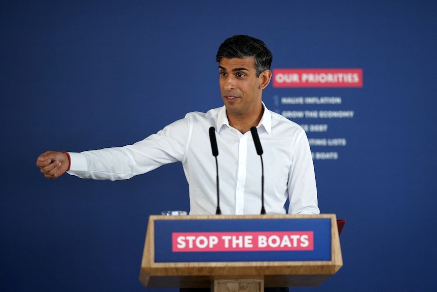 A man speaking behind a lecturn that has a sign on it that says "stop the boats"