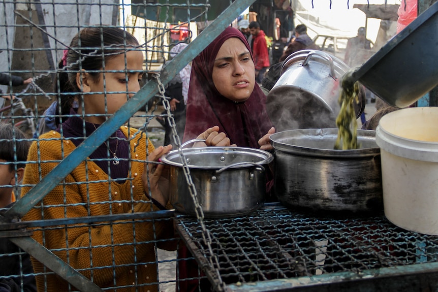 A young woman and a girl queue with metal pots to receive a free meal