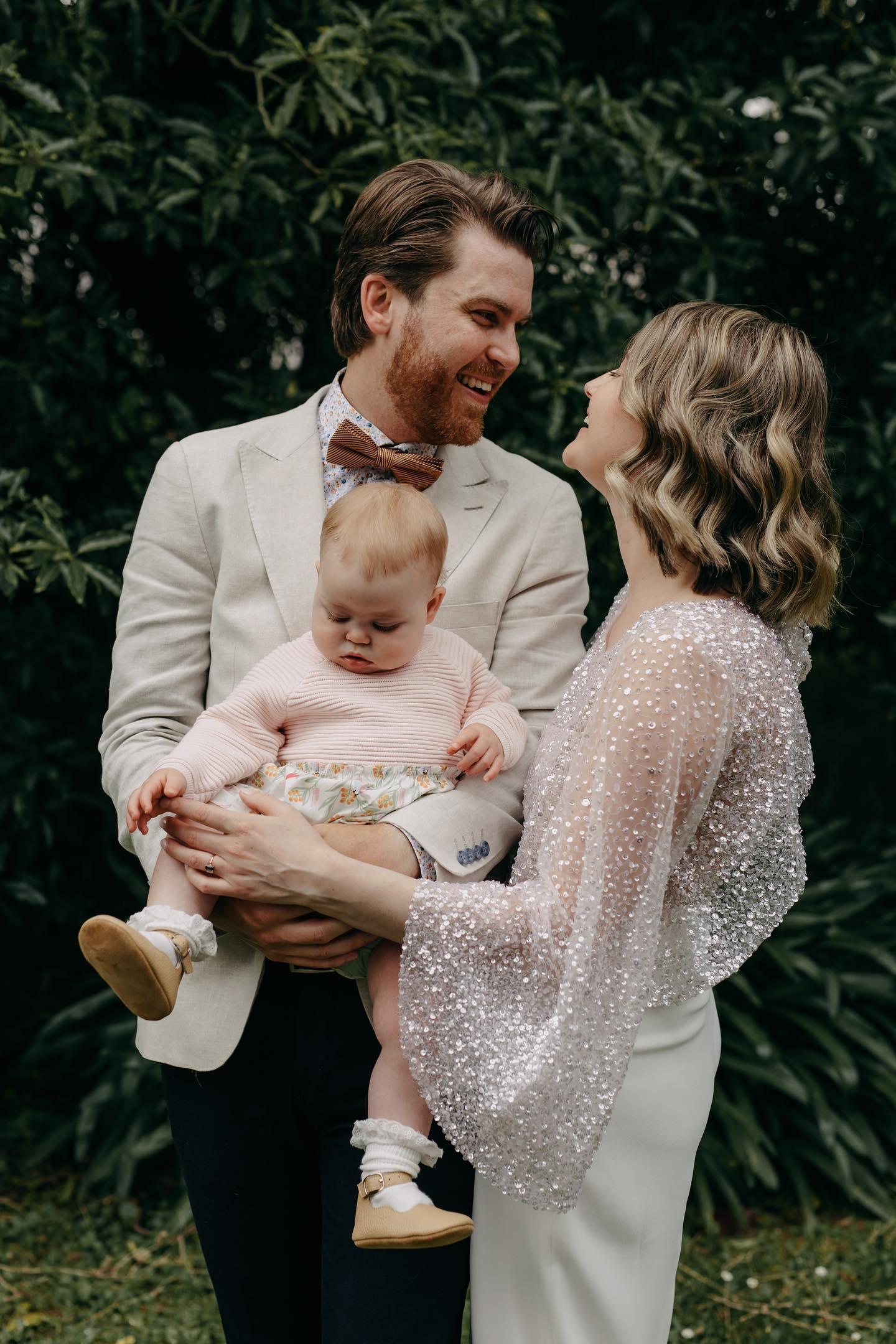 A portrait family photo showing smiling husband and wife holding a baby in their arms.